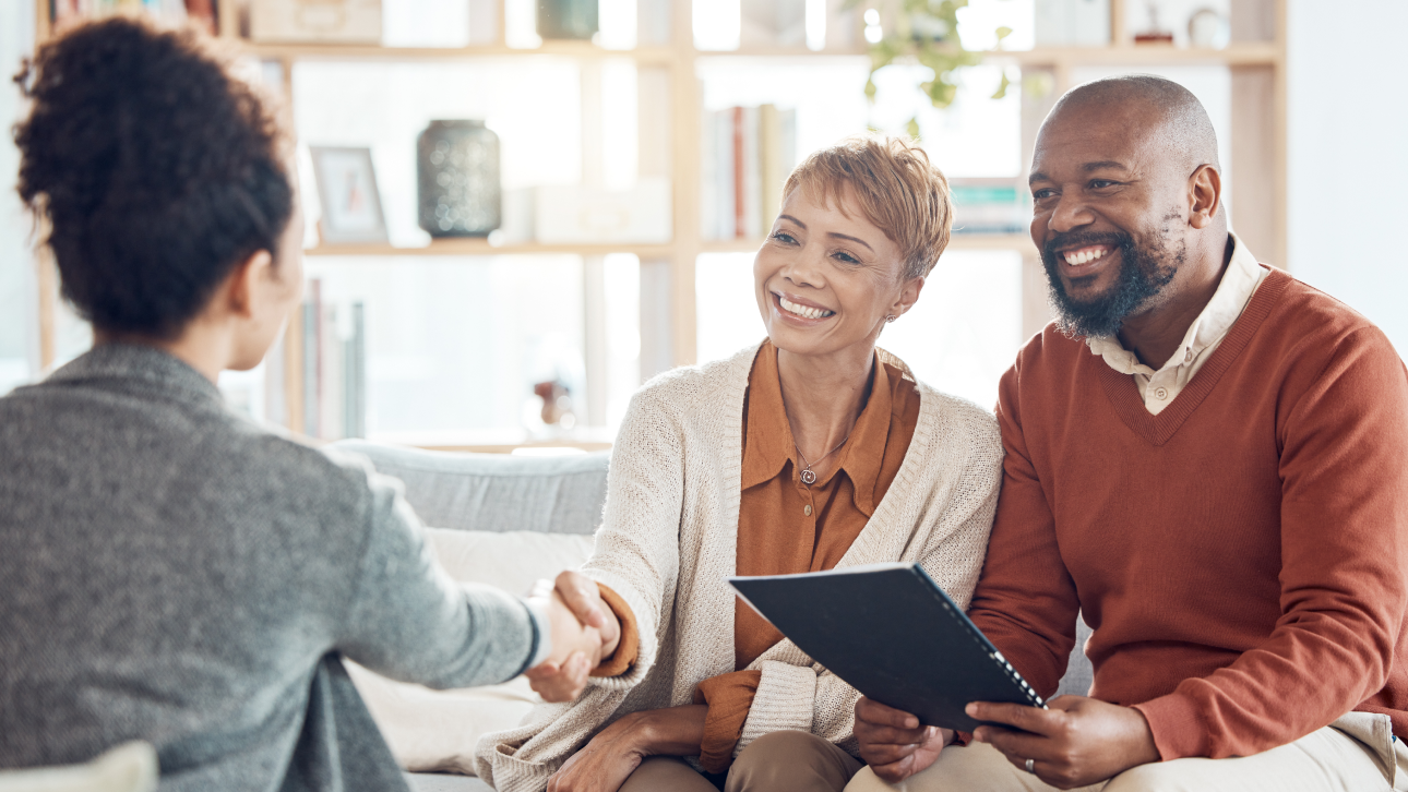 A smiling couple siting on couch shaking hands with woman seated across from them