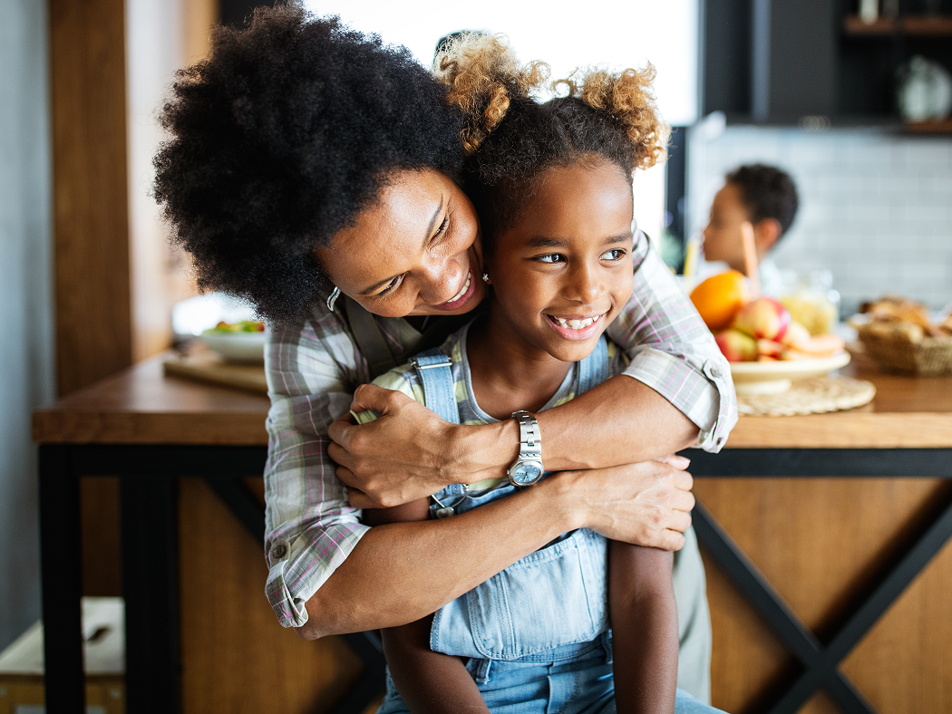 Smiling woman embraces a young girl from behind, both standing in a kitchen with fruit and food visible on the counter in the background