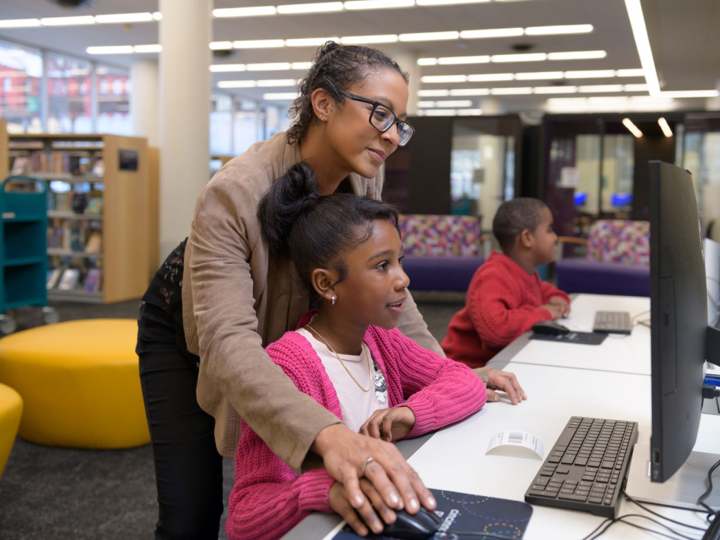 Woman helps a young girl use a desktop computer in a library, while a boy in the background works at another computer.