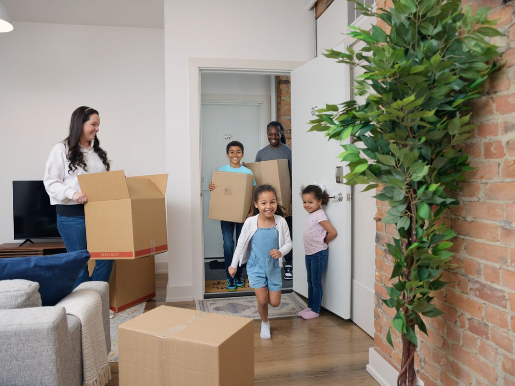 Smiling family moves into a new home, carrying cardboard boxes, as two young girls run inside